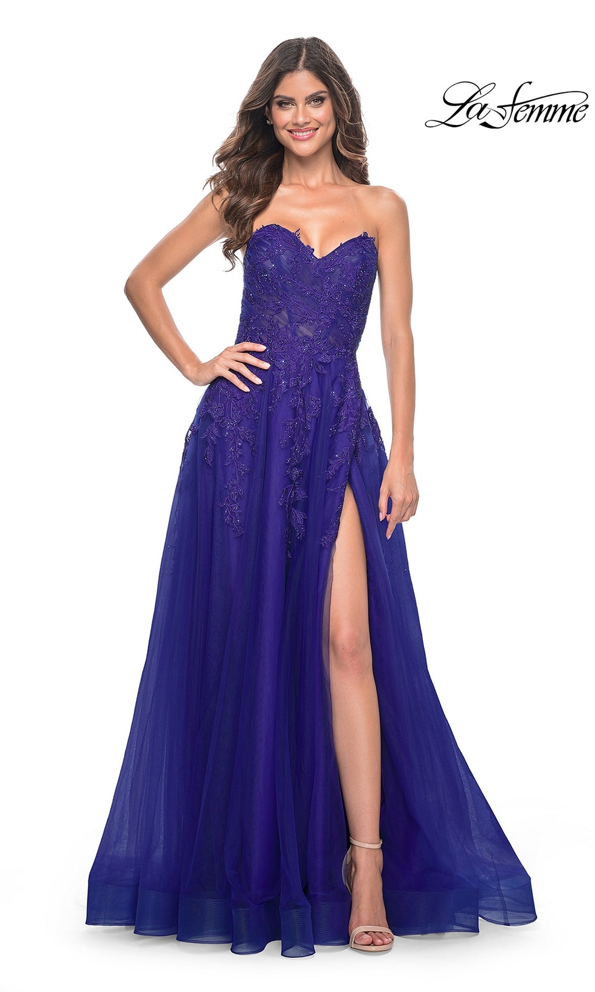 A woman poses confidently with one hand on her hip in the La Femme Lace-Up Long Sweetheart Prom Dress 32304, a strapless royal blue gown featuring lace details and a high thigh slit.